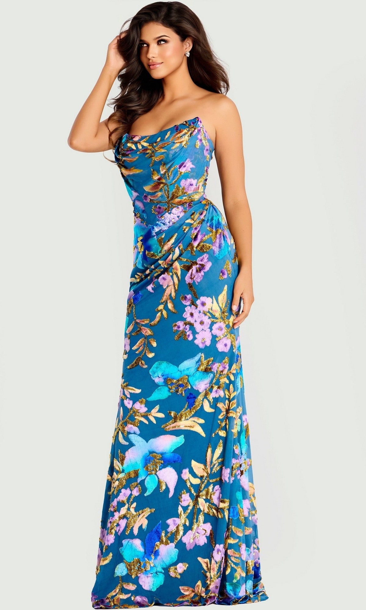 A woman stands against a light background wearing the Jovani 47654, a long blue prom dress with a floral print and draped neckline.